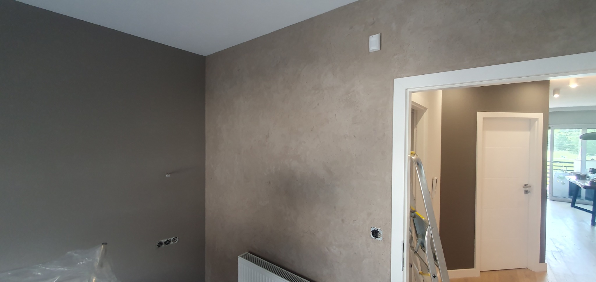 Ściana w odcieniach szarości z dekoracyjną fakturą, widoczne gniazdka elektryczne przed montażem, grzejnik i drabina malarska przy futrynie drzwi.
