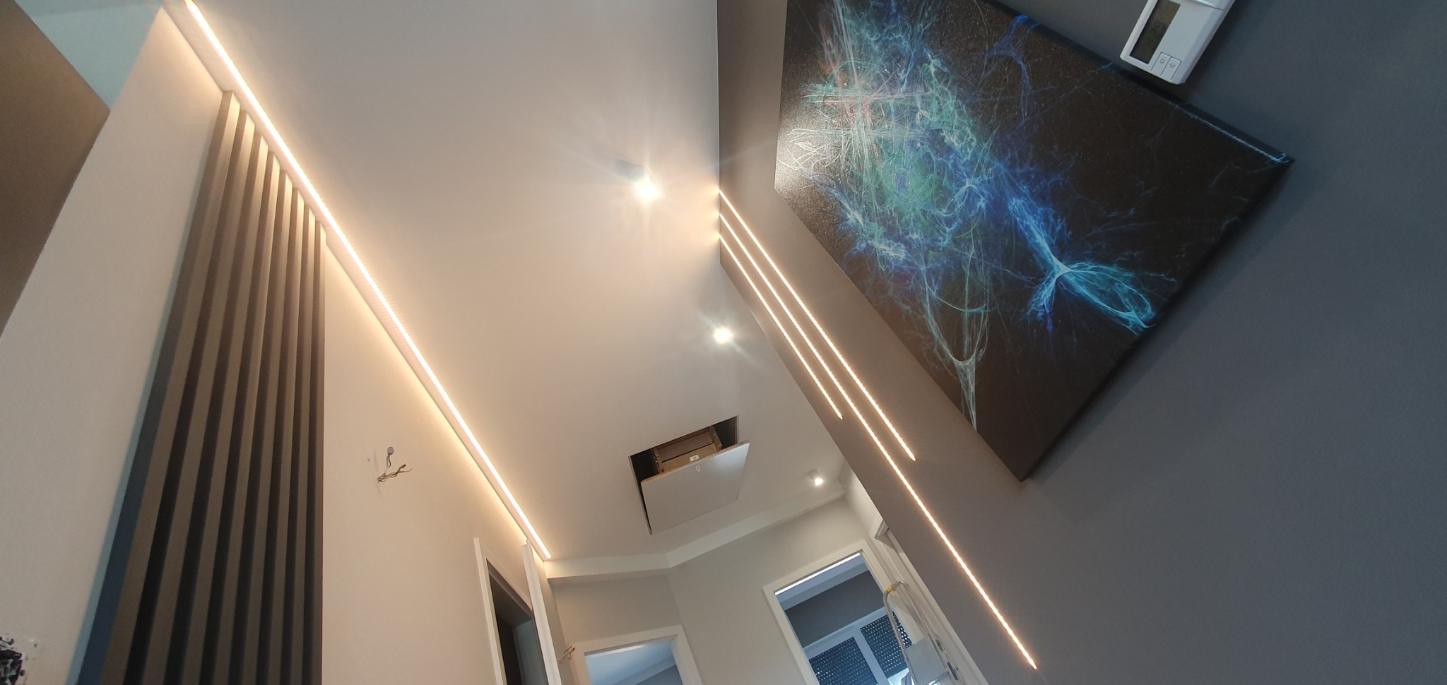 Nowoczesne oświetlenie LED wzdłuż sufitu i ściany, dekoracyjny panel ścienny z pionowych listew, abstrakcyjny obraz na ścianie, widok na korytarz z otwartymi drzwiami.