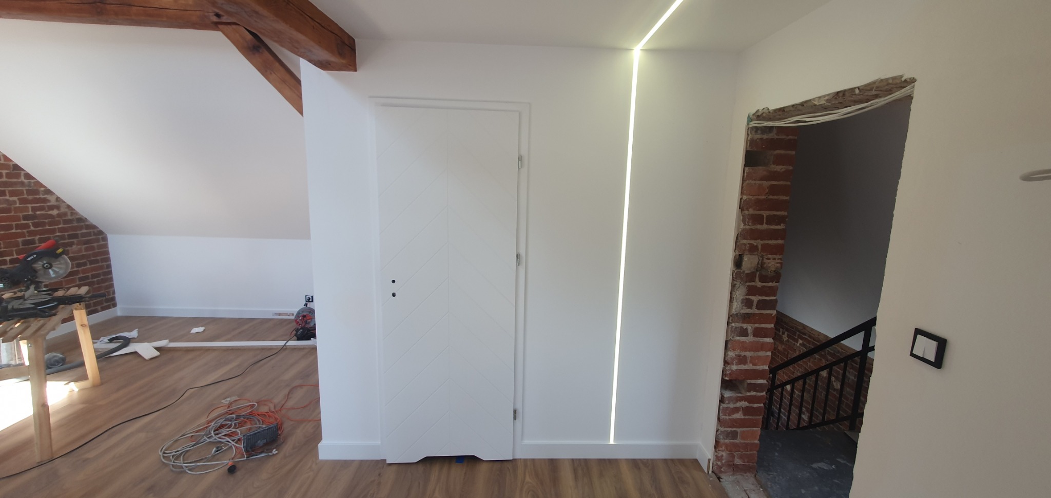 Poddasze w trakcie prac wykończeniowych: biała ściana z pionowym oświetleniem LED, drzwi z dekoracyjnym wzorem, fragment ściany z cegły z widocznymi przewodami i schodami w tle, oraz narzędzia...