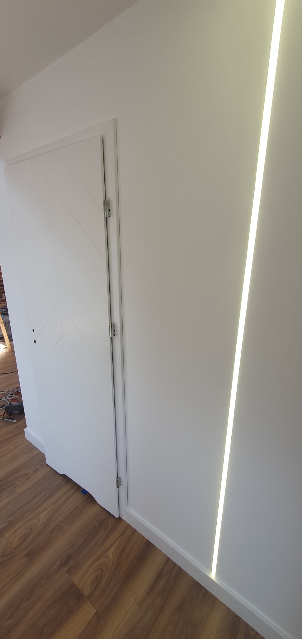 Biała, ukryta w ścianie pionowa listwa LED, oświetlająca fragment białej ściany z drzwiami o wzorze jodełki, wykończona białą listwą przypodłogową.