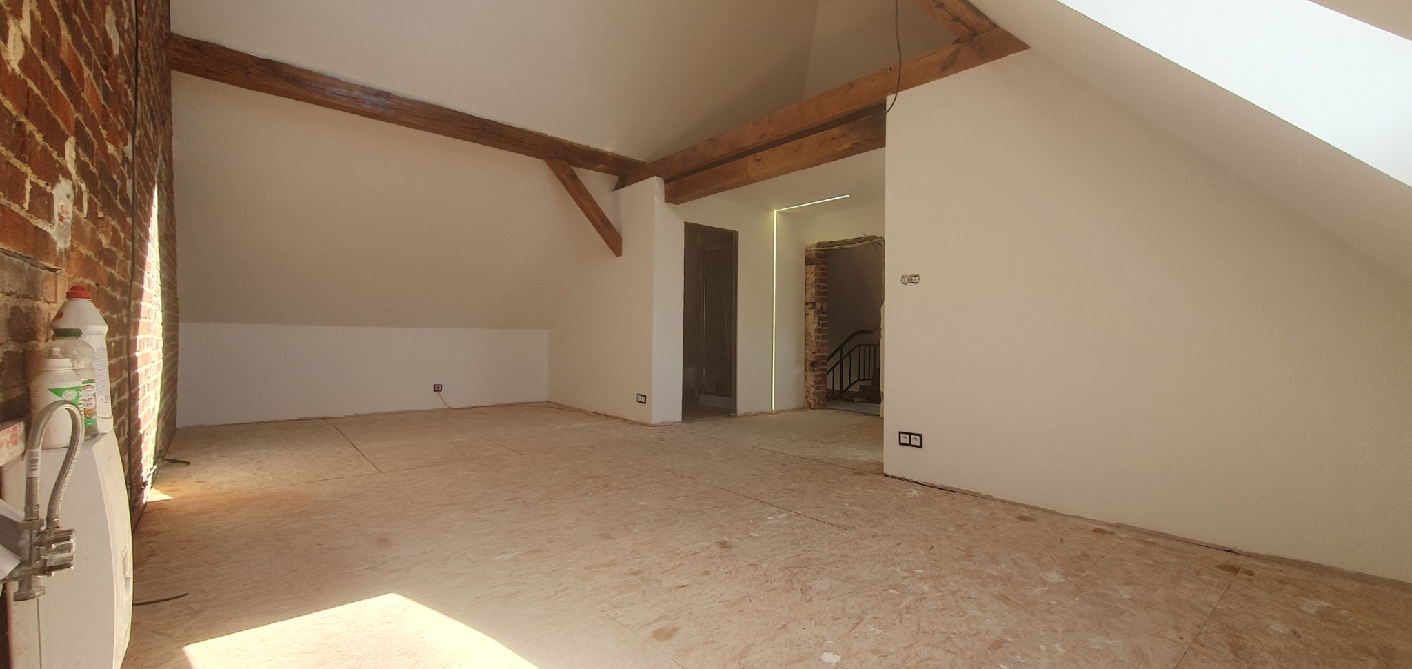 Poddasze w trakcie adaptacji: widoczne drewniane belki konstrukcyjne, ściany przygotowane pod malowanie, surowa podłoga z płyt OSB, instalacja elektryczna w ścianach, fragment ściany z cegły...