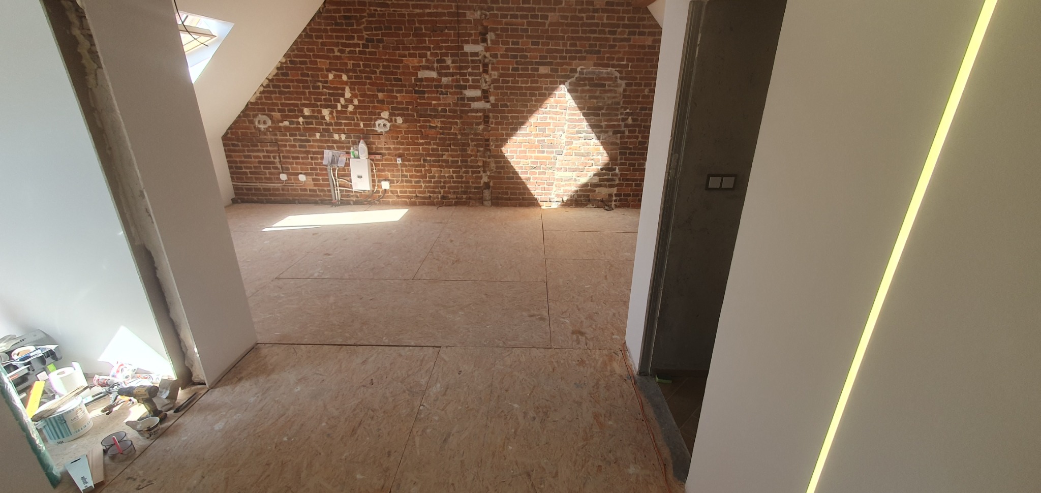Poddasze w trakcie prac wykończeniowych: surowa ceglana ściana, drewniana podłoga z płyt OSB, przygotowane ściany działowe, narzędzia i materiały budowlane w rogu.