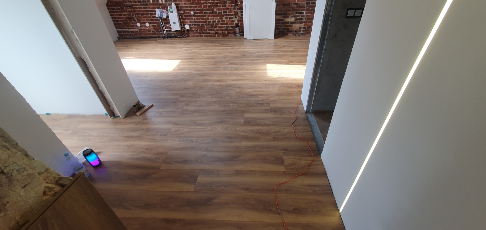 Nowo położona podłoga laminowana w jasnym odcieniu drewna, widoczne fragmenty ścian z surowej cegły i świeżo pomalowane na biało ściany z oświetleniem LED, pomarańczowy kabel elektryczny leżący...