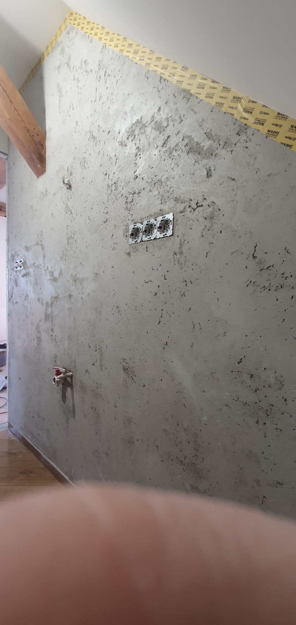 Ściana w trakcie nakładania dekoracyjnej, szarej gładzi z widocznymi puszkami elektrycznymi bez osprzętu, zabezpieczona taśmą malarską przy suficie i elementem drewnianym poddasza.