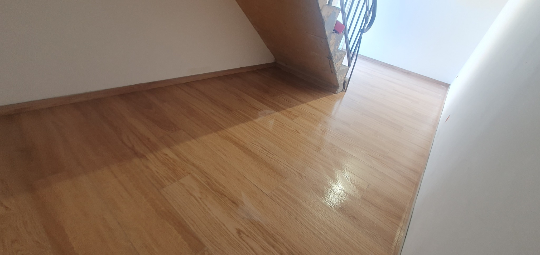 Świeżo położona, błyszcząca podłoga laminowana w odcieniach drewna, widok spod schodów, białe ściany z widocznymi śladami po remoncie.
