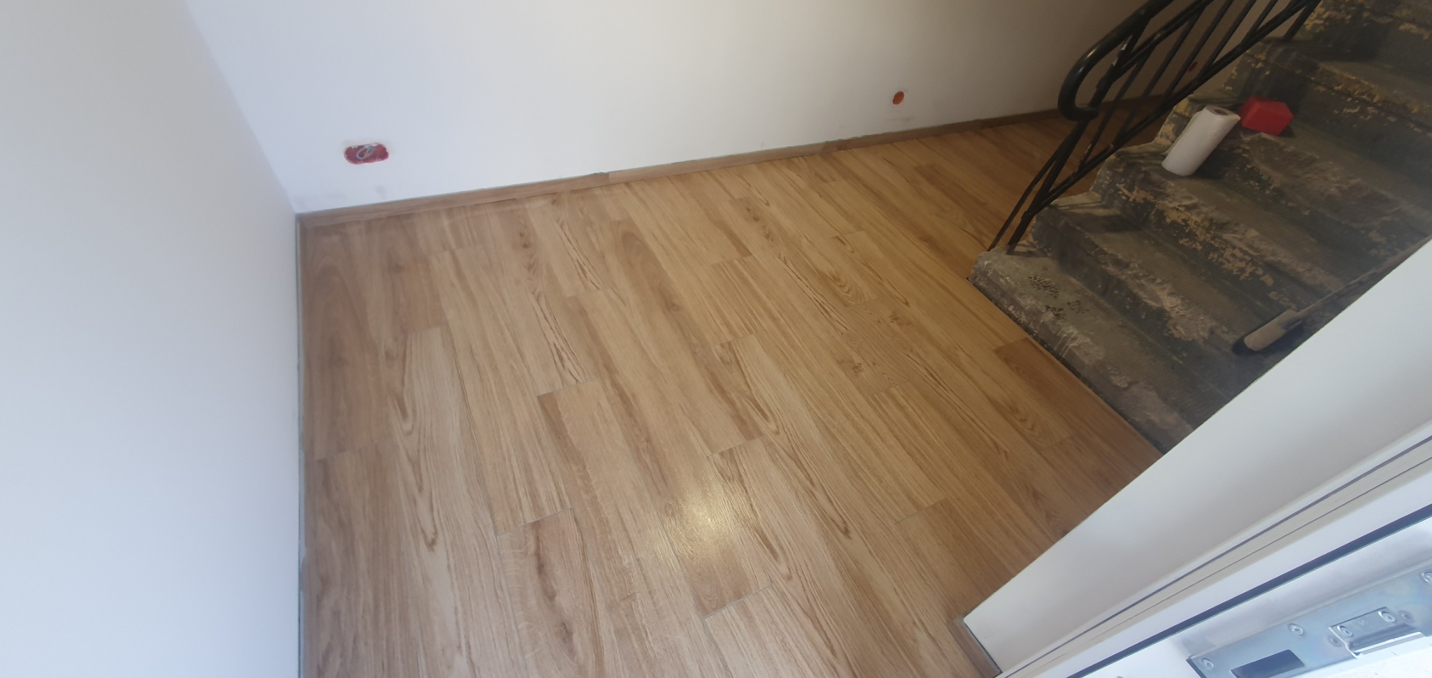 Ułożona podłoga panelowa w korytarzu, widoczne schody z czarną balustradą i białe ściany z wystającymi przewodami elektrycznymi.