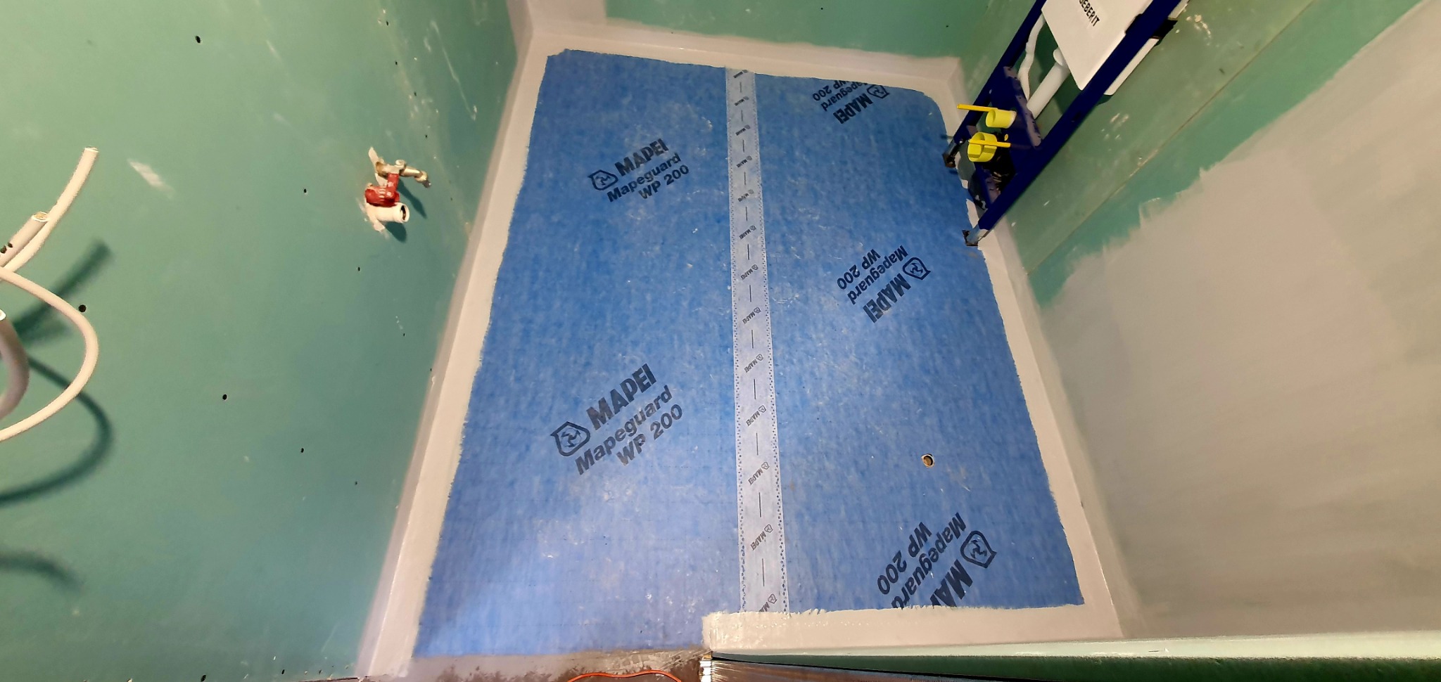 Przygotowanie podłoża pod prysznic z użyciem hydroizolacji Mapeguard WP 200, widoczna instalacja Geberit i surowa ściana z wyprowadzonym przyłączem wodnym.