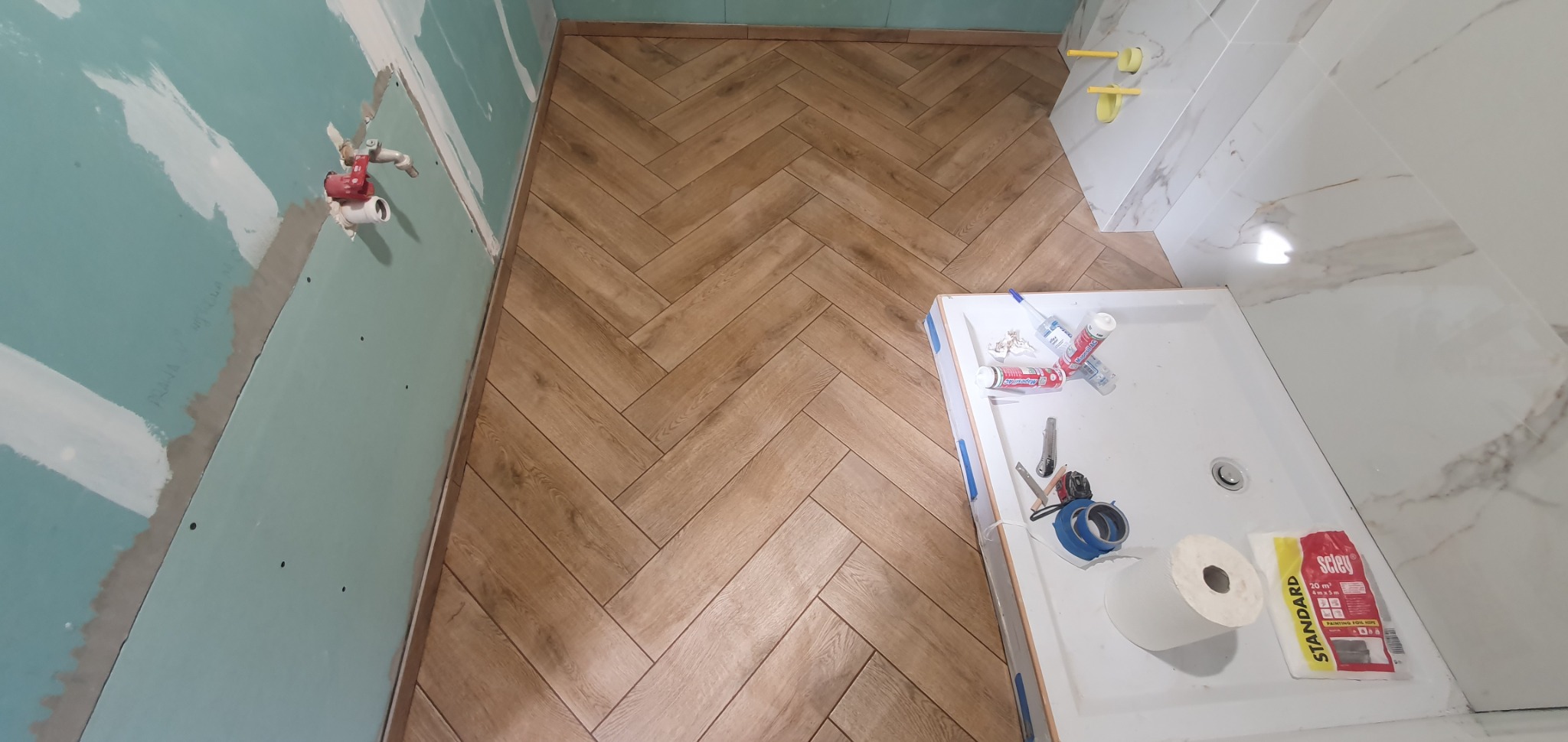 Montaż brązowych płytek podłogowych w jodełkę w trakcie wykańczania łazienki, widoczne narzędzia, nieotynkowana ściana z płyt karton-gips i biały brodzik.