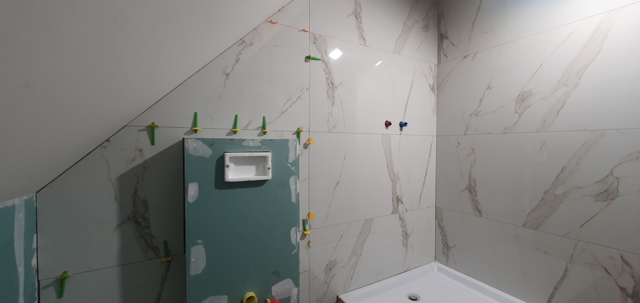 Ściany łazienki w trakcie wykańczania, widoczne płytki imitujące marmur, poziomowanie z użyciem klinów, zabudowa stelaża WC z płyt karton-gips.