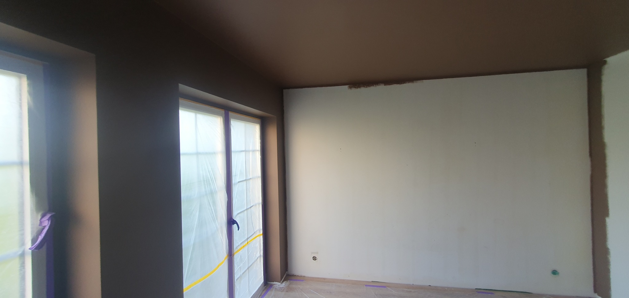 Pokój po malowaniu ścian na biało i brązowo, okna zabezpieczone folią malarską. Widoczne ślady taśmy malarskiej na podłodze.
