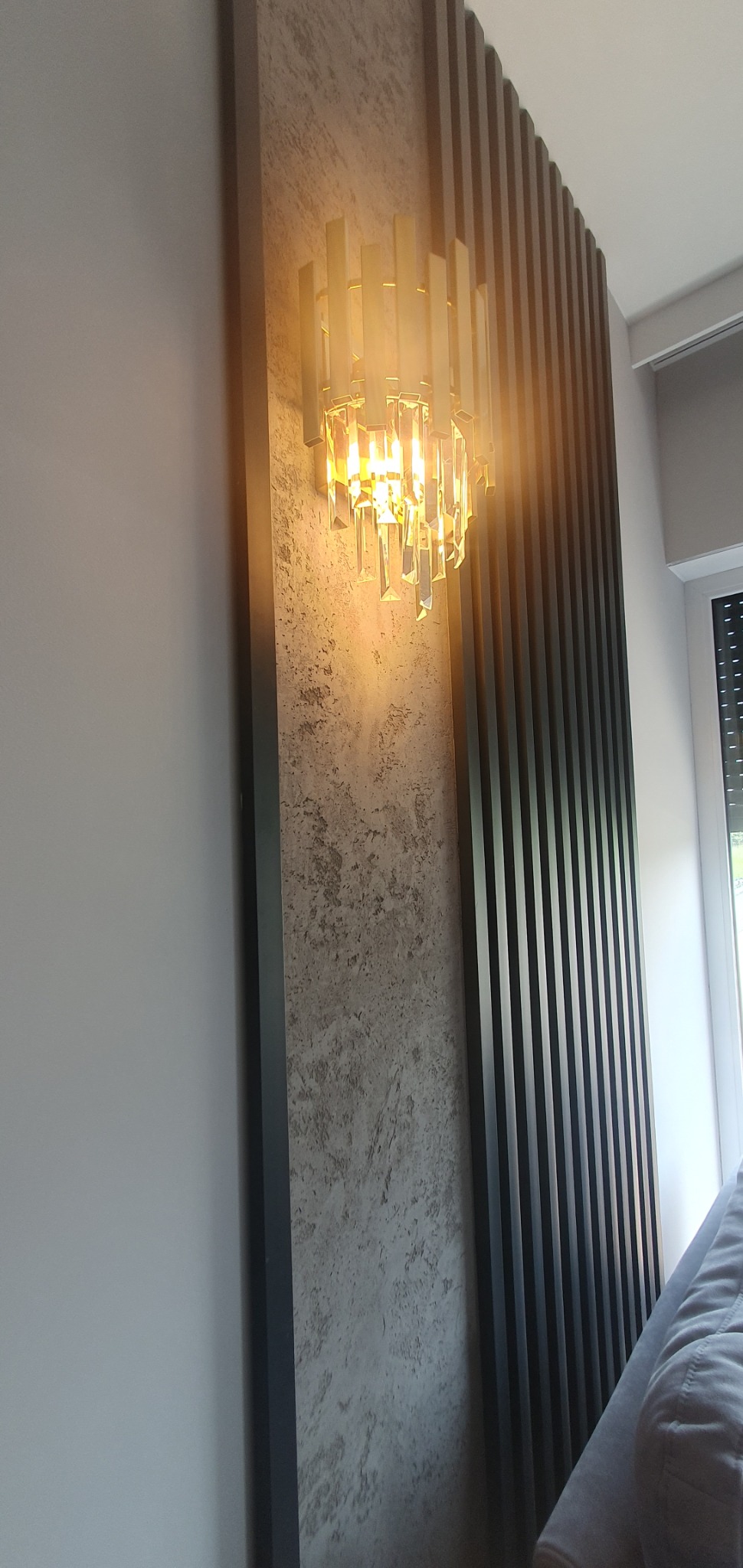 Nowoczesna ściana z dekoracyjnymi lamelami w odcieniach szarości, oświetlona kryształowym kinkietem. Element wystroju wnętrza, widoczne fragmenty okna i kanapy.