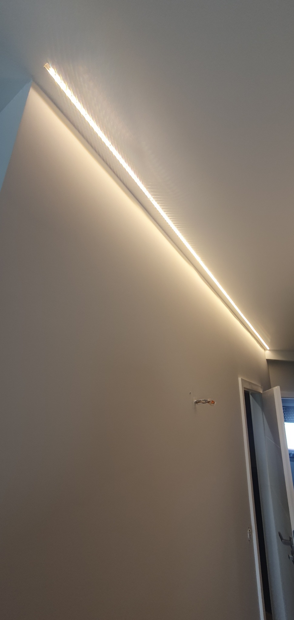 Nowoczesne oświetlenie LED wzdłuż sufitu, świecące ciepłym światłem, na tle gładkiej, szarej ściany. Widoczne fragmenty drzwi i instalacji elektrycznej.