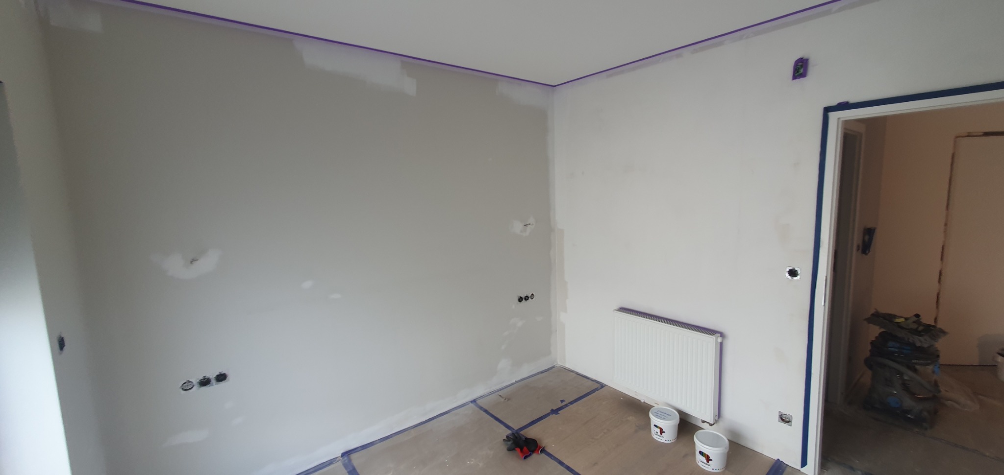 Pokój w trakcie szpachlowania i przygotowania do malowania, zabezpieczone listwy i podłoga taśmą malarską, widoczne puszki z farbą i narzędzia.