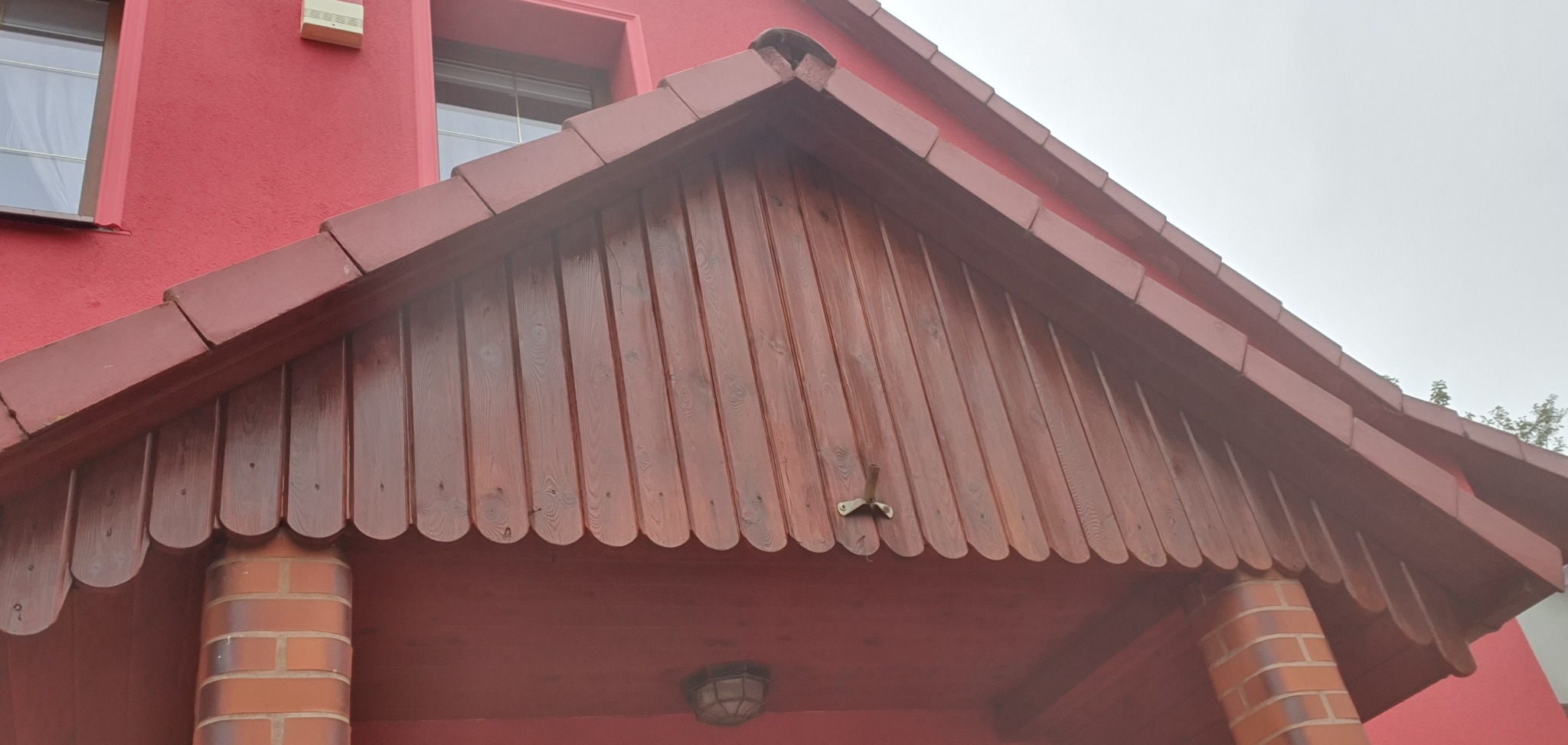 Czerwony dom z drewnianą, lakierowaną podbitką dachową i ceglanymi filarami wejścia, widziany od dołu pod kątem. Widoczne okno i alarm.