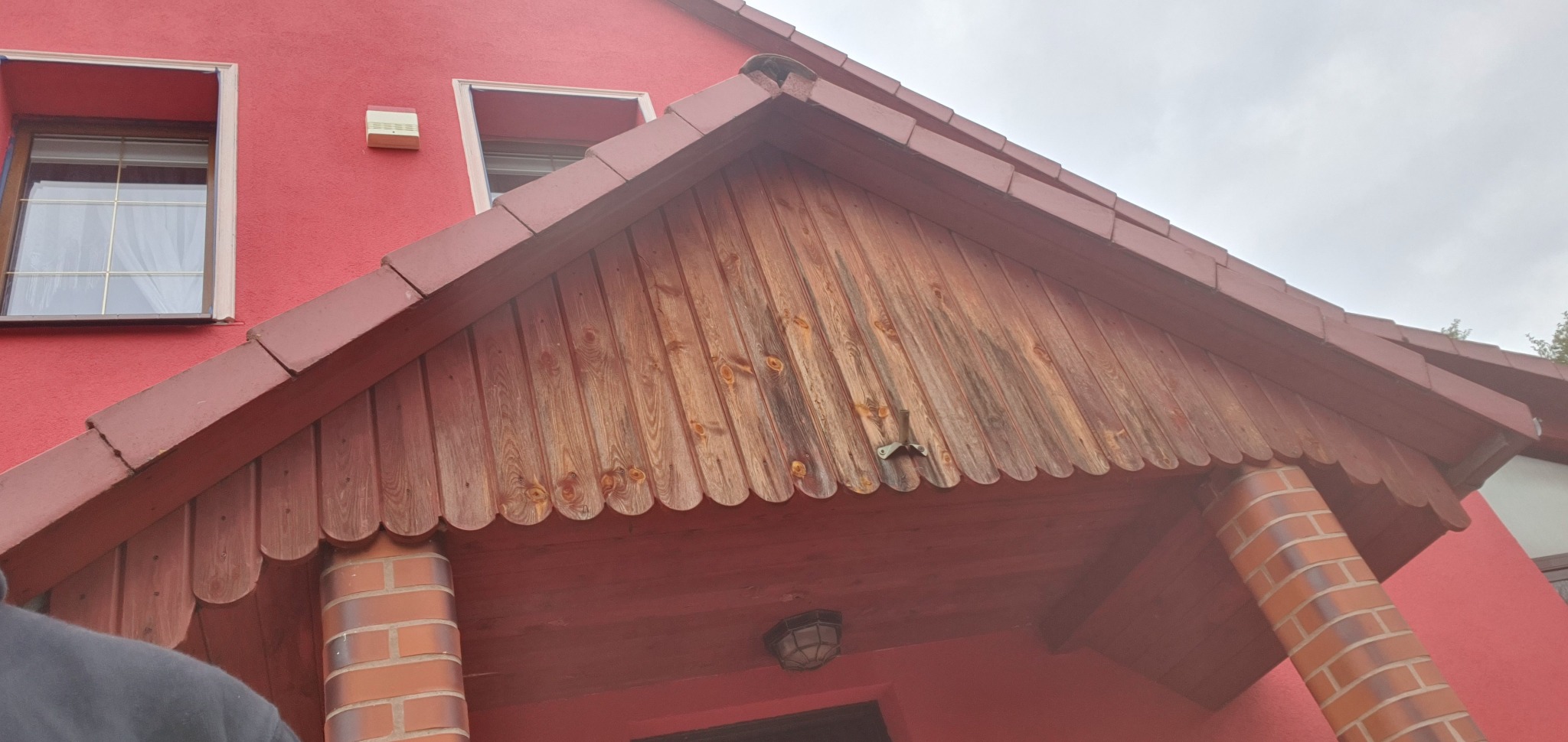 Czerwony dom z drewnianym zadaszeniem nad wejściem, wspartym na ceglanych kolumnach. Widoczne okna i fragment elewacji. Zadaszenie z widocznymi śladami użytkowania.