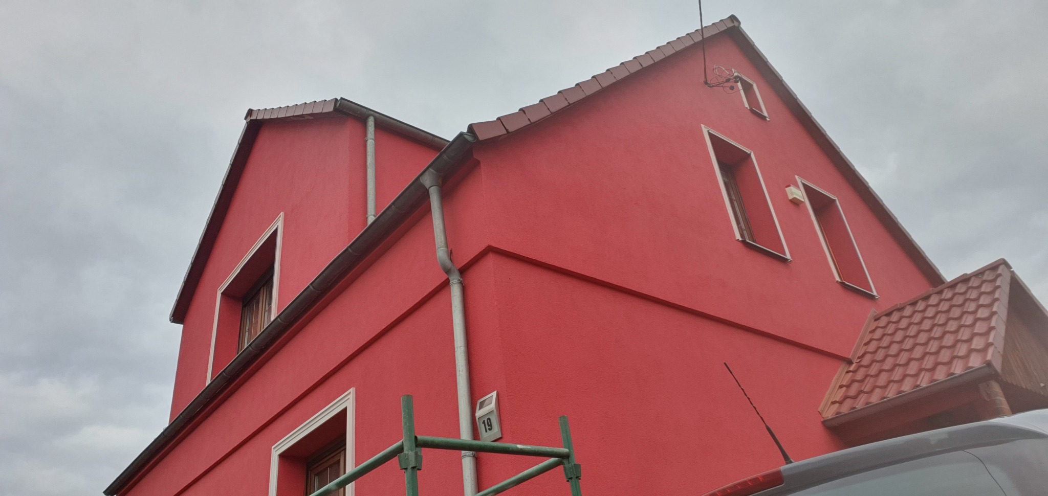Czerwony dom po renowacji elewacji, widok z dołu, z fragmentem rusztowania i samochodu w kadrze. Widoczne okna z białymi ramami i brązowy dach.