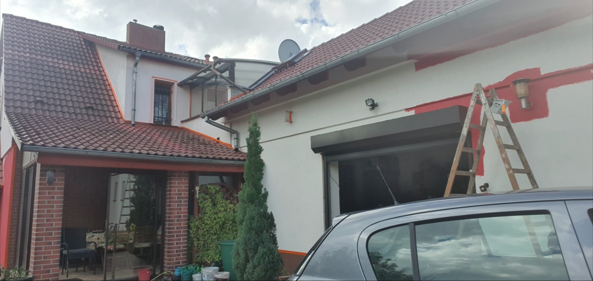 Malowanie elewacji domu w trakcie: biała ściana z czerwonymi fragmentami, drabina oparta o ścianę, widoczne puszki z farbą i fragment samochodu na pierwszym planie.