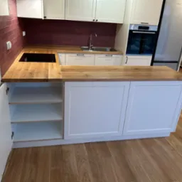 Nowoczesna kuchnia z białymi szafkami, drewnianym blatem i bordowymi płytkami na ścianie, widoczna płyta indukcyjna, zlew i otwarte szafki z p&oacute;łkami.