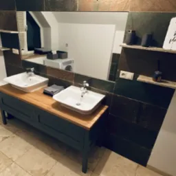 Łazienka z dwiema białymi umywalkami na drewnianym blacie, ciemnoszara szafka pod umywalkami, lustro z dekoracyjną ramą z płytek, p&oacute;łki z akcesoriami i dekoracją 'This is my happy place', pralka...