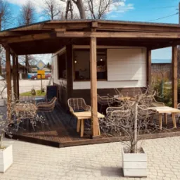 Zadaszony taras drewniany z rattanowymi krzesłami i stolikami, otoczony kostką brukową i donicami z małymi drzewkami.