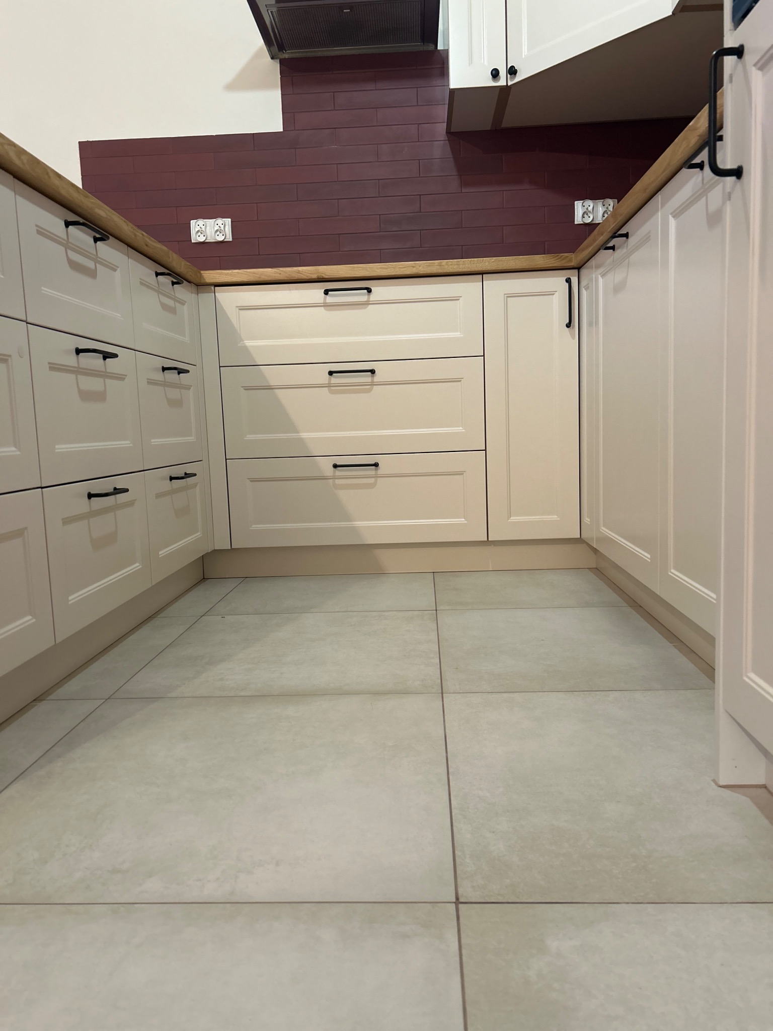 Jasna zabudowa kuchenna z ciemnymi uchwytami i drewnianym blatem w Wygrynach, uzupełniona bordową cegłą na ścianie i szarymi płytkami na podłodze, widok z dołu.