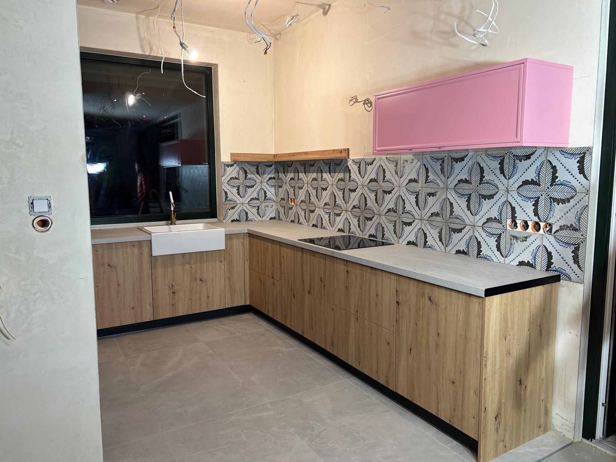 Wnętrze kuchni w trakcie montażu, widoczne drewniane szafki dolne z szarym blatem, dekoracyjne płytki na ścianie oraz różowa szafka wisząca, surowy stan ścian i wystające kable.