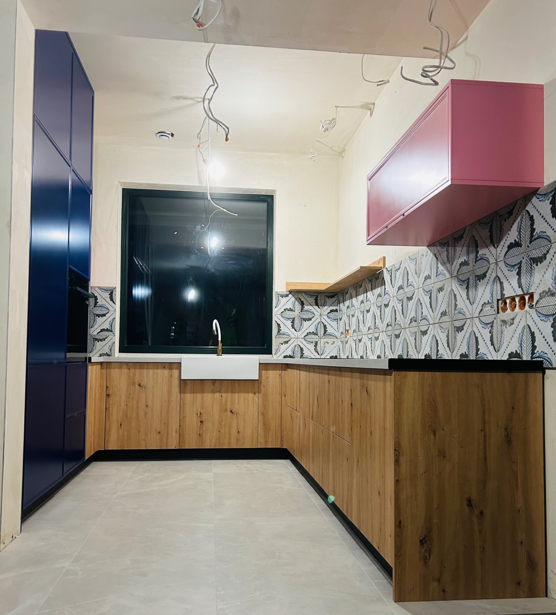 Wnętrze kuchni w trakcie montażu: drewniane szafki dolne z czarnymi listwami, granatowa wysoka zabudowa, różowa szafka wisząca, dekoracyjne płytki nad blatem, widoczne przewody elektryczne.