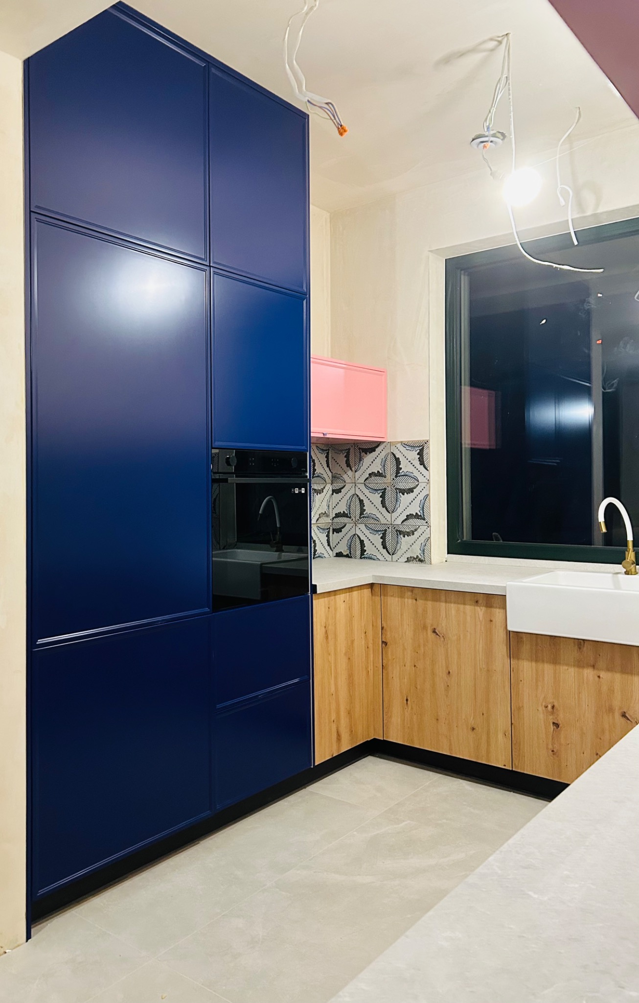 Niebieska zabudowa kuchenna z piekarnikiem w trakcie montażu, widoczne kable oświetleniowe, drewniane szafki dolne i różowa szafka wisząca nad wzorzystymi płytkami.