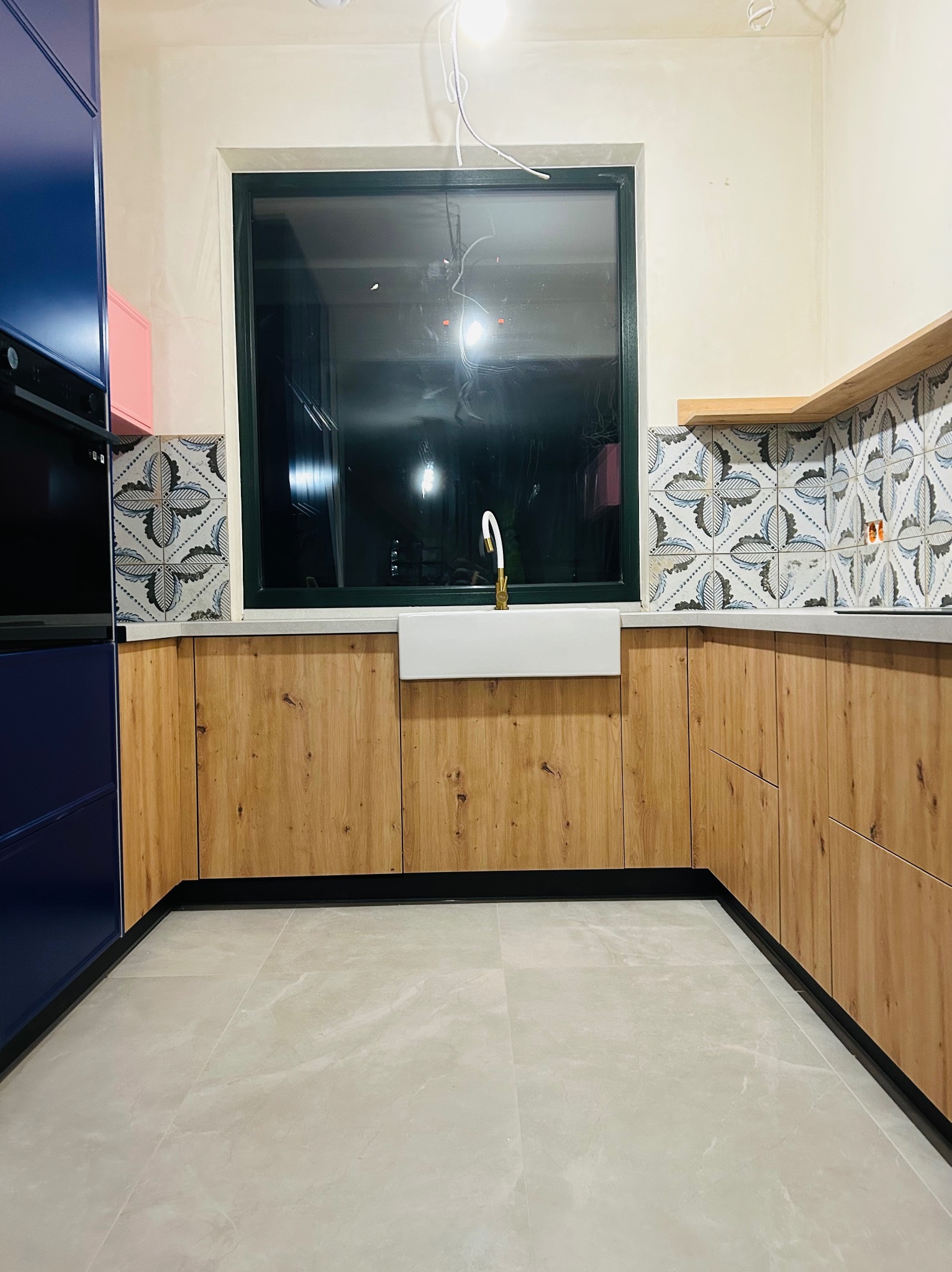 Nowoczesna aranżacja kuchni z drewnianymi szafkami, wzorzystymi płytkami nad blatem, białą ceramiczną umywalką i złotą baterią, widok na okno.