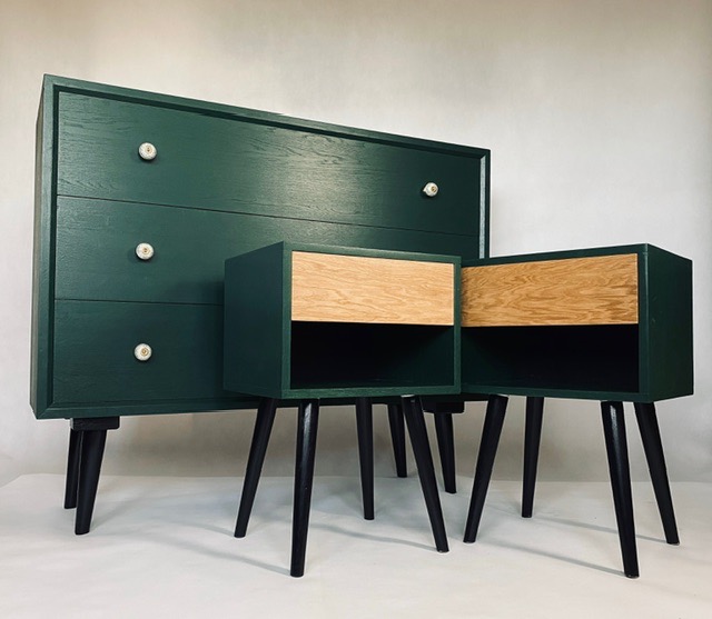 Komoda z trzema szufladami w kolorze butelkowej zieleni z okrągłymi, kremowymi uchwytami, obok dwie szafki nocne z jasnymi, drewnianymi szufladami i czarnymi nóżkami w stylu retro na białym tle.