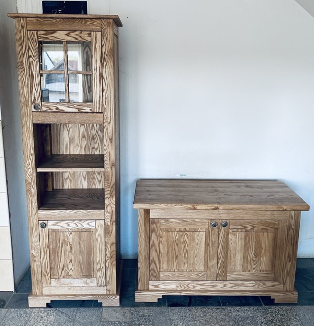 Dwa meble z jasnego drewna o wyraźnym usłojeniu: wysoki regał z przeszkloną szafką na górze i niską szafką z podwójnymi drzwiczkami, stojące na ciemnej posadzce przy białej ścianie.