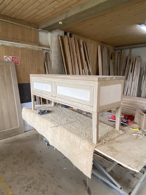 Niedokończona szafka z jasnego drewna z białymi panelami, umieszczona na stole warsztatowym pokrytym wiórami, w tle deski i ściany warsztatu.