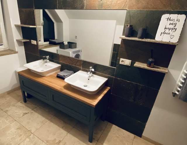 Łazienka z dwiema białymi umywalkami na drewnianym blacie, ciemnoszara szafka pod umywalkami, lustro z dekoracyjną ramą z płytek, półki z akcesoriami i dekoracją 'This is my happy place', pralka...