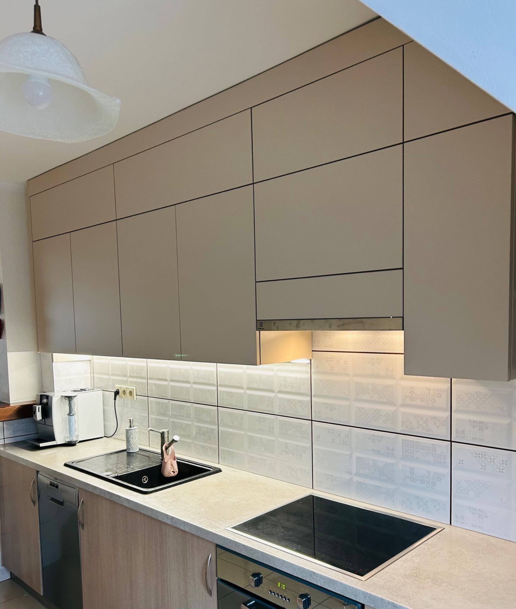 Nowoczesna kuchnia z beżowymi szafkami górnymi z czarnymi obramowaniami, białymi płytkami z delikatnym wzorem, czarnym zlewem i płytą indukcyjną oraz wiszącą lampą.