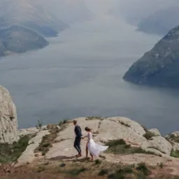 Para młoda wędruje po skalistym szczycie z widokiem na fiord. Panna młoda w zwiewnej sukni, pan młody w granatowym garniturze.