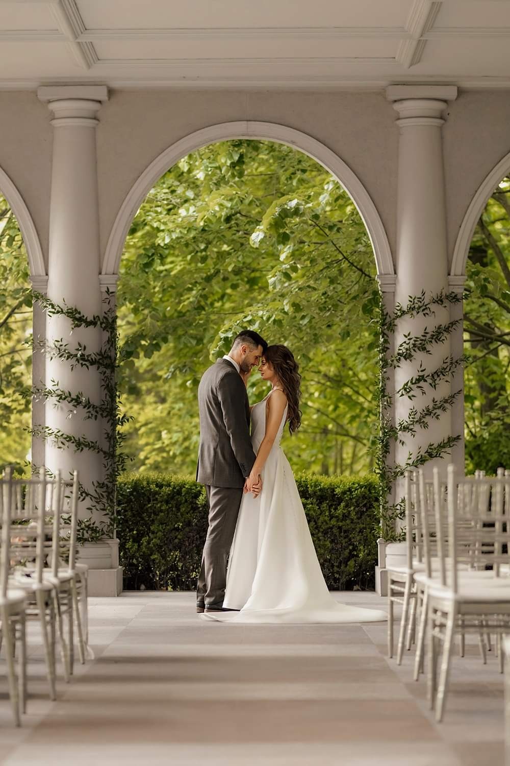 Para młoda trzymająca się za ręce pod ozdobnym łukiem z kolumnami porośniętymi bluszczem, w tle zielona roślinność i rzędy krzeseł dla gości.