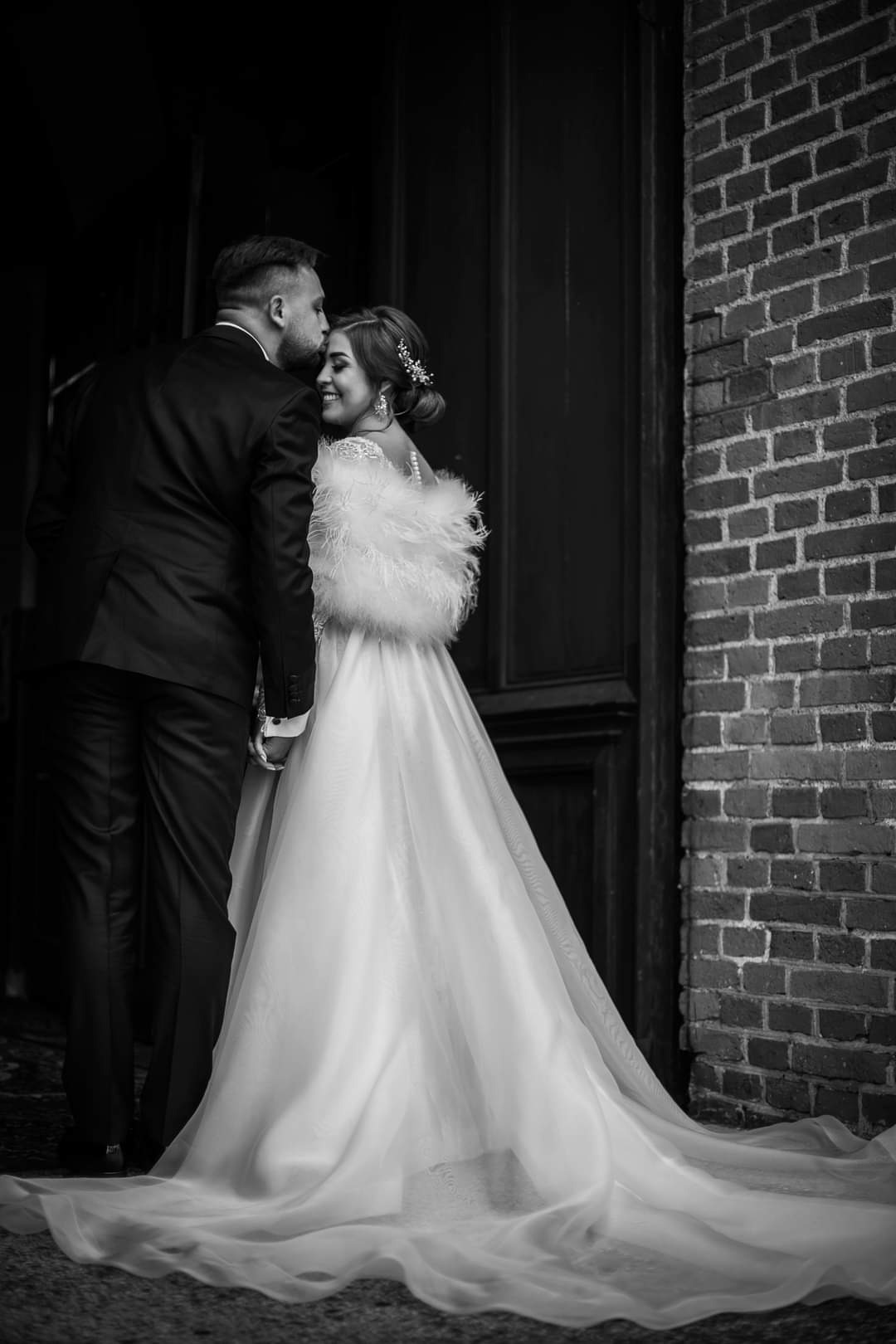 Czarno-białe zdjęcie: Panna młoda w sukni z piórami i upięciem we włosach uśmiecha się, gdy pan młody całuje ją w czoło na tle ceglanej ściany i ciemnych drzwi.