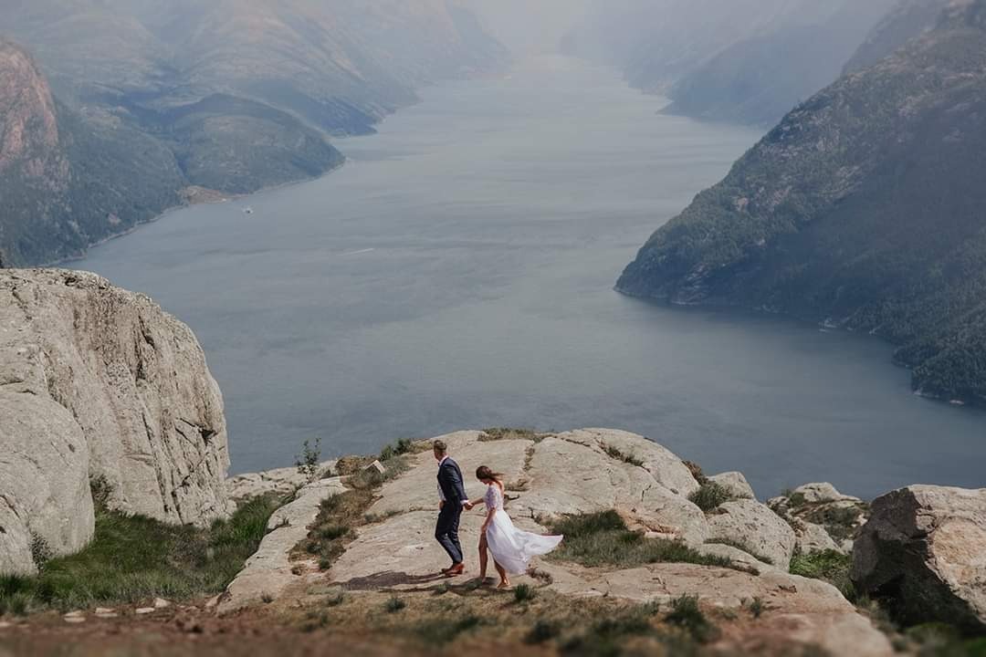 Para młoda wędruje po skalistym szczycie z widokiem na fiord. Panna młoda w zwiewnej sukni, pan młody w granatowym garniturze.