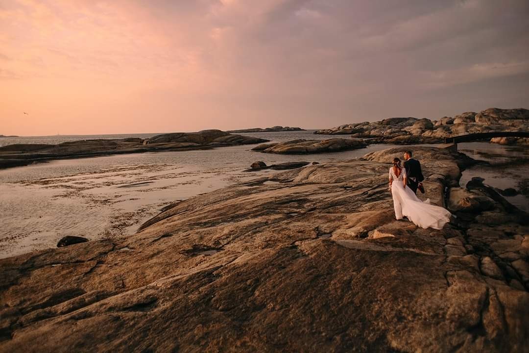 Para młoda spaceruje po skalistym wybrzeżu o zachodzie słońca, suknia panny młodej powiewa na wietrze.
