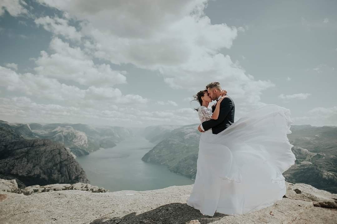 Para młoda całuje się na tle malowniczego fiordu i skalistych gór w Norwegii, suknia powiewa na wietrze.