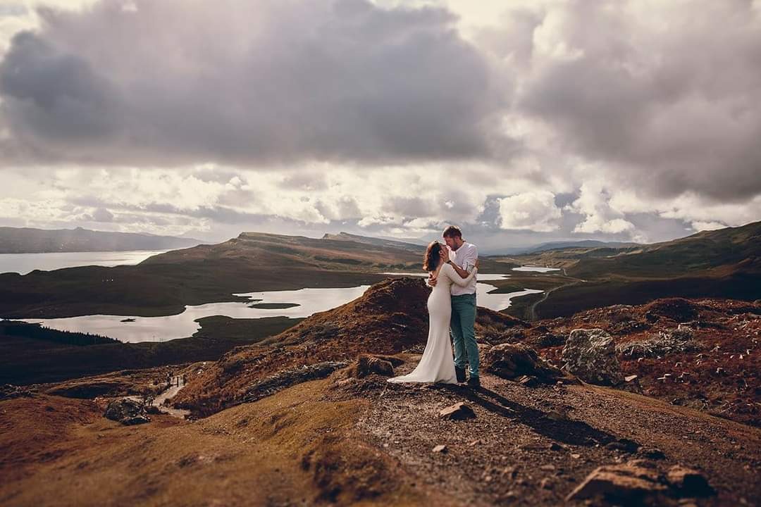 Para w objęciach na szczycie wzgórza z widokiem na jeziora i góry pod pochmurnym niebem. Panna młoda w długiej białej sukni, pan młody w białej koszuli i jeansach.