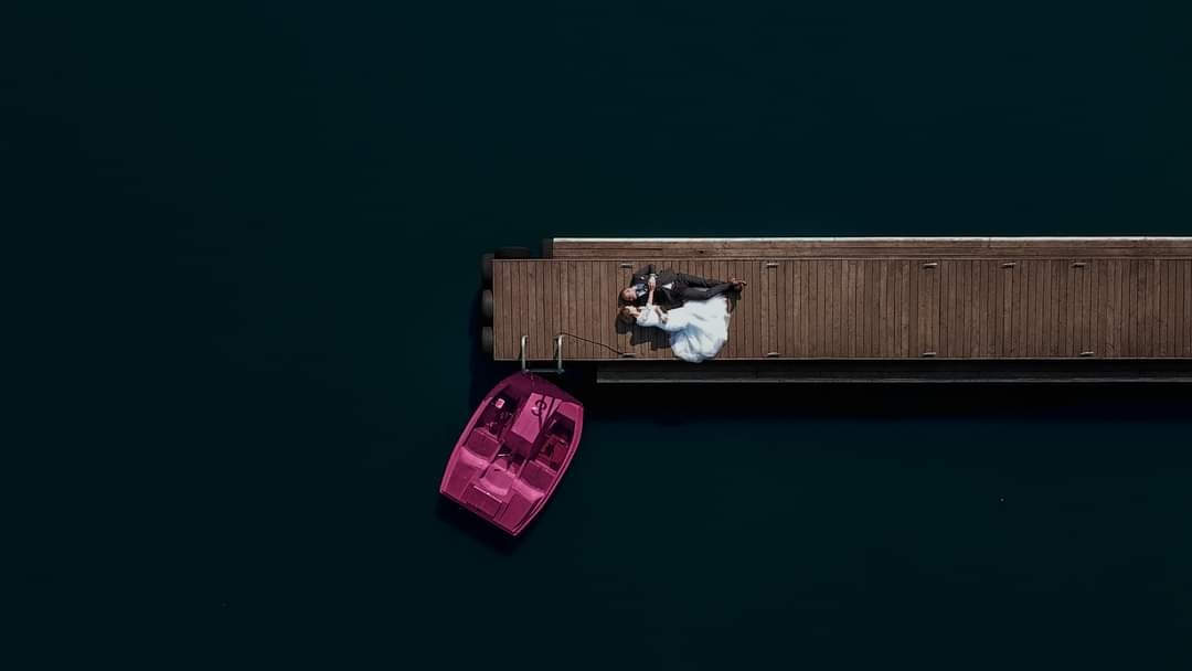 Para młoda leżąca na drewnianym molo nad ciemną wodą, obok różowa łódka; widok z góry.