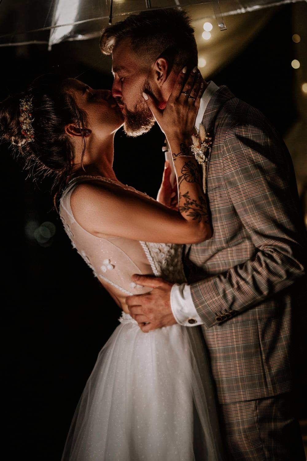 Pocałunek nowożeńców pod przezroczystym parasolem, panna młoda w sukni z delikatnymi kwiatowymi aplikacjami na plecach, pan młody w brązowym garniturze w kratę, ciemne tło z rozproszonymi światłami.