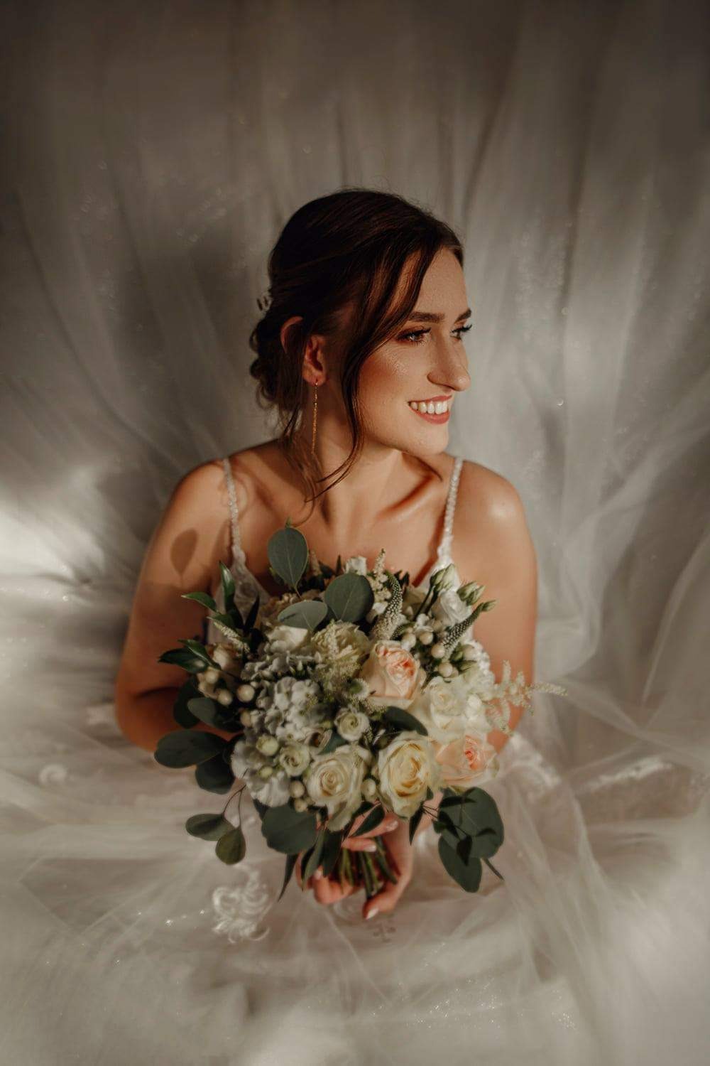 Uśmiechnięta panna młoda w białej sukni, trzymająca bukiet ślubny z białych i różowych róż oraz zielonych liści eukaliptusa na tle delikatnej, białej tkaniny.