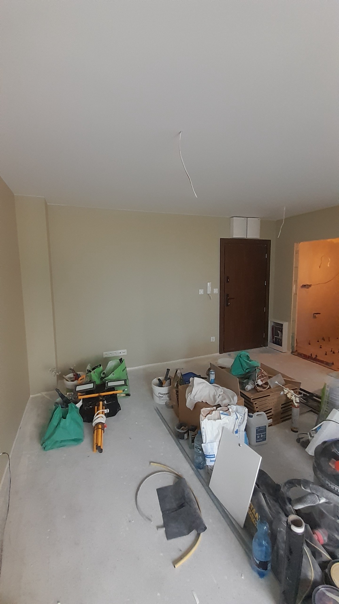 Wnętrze w trakcie prac wykończeniowych: świeżo pomalowane ściany w odcieniach beżu, surowy beton na podłodze, widoczne narzędzia i materiały budowlane, w tle ciemne drzwi i otwór do kolejnego...