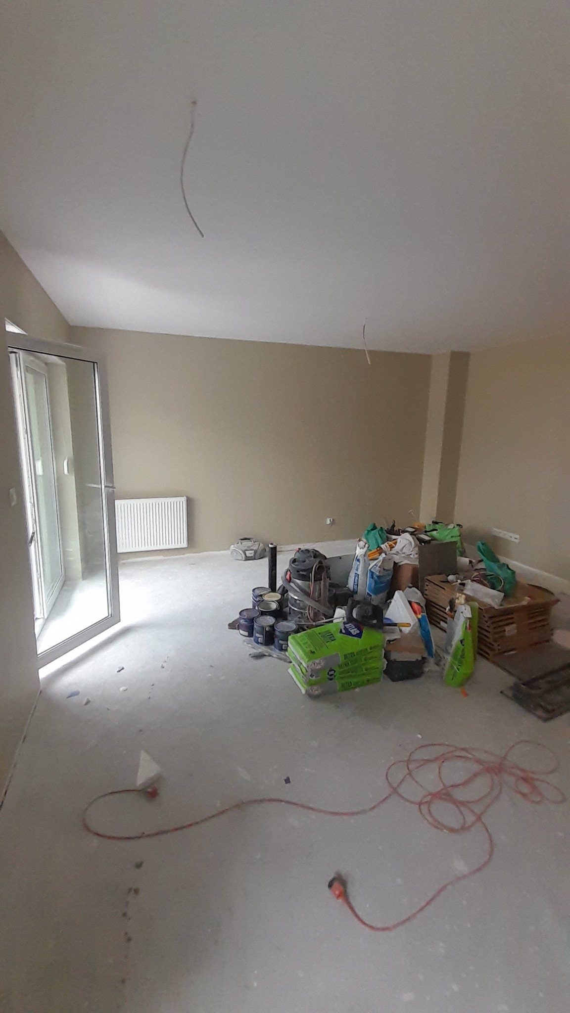 Pokój w trakcie prac wykończeniowych: widoczne puszki farb, worki z zaprawą, odkurzacz przemysłowy, grzejnik na ścianie i przewody elektryczne na podłodze.