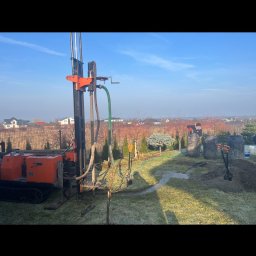 Wiercenie Studni Tadeusz Sadłowski - Wiercenie studni głębinowej w ogrodzie za pomocą specjalistycznej maszyny wiertniczej. Widoczne czarne zbiorniki na wodę i wykopana ziemia oraz pracownik w tle.