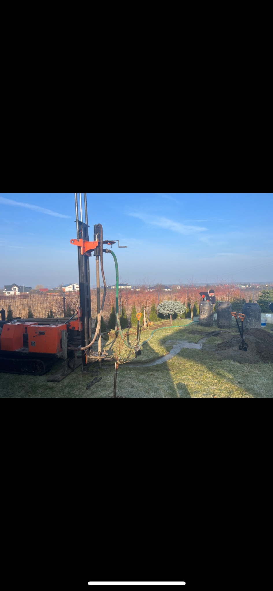 Wiercenie studni głębinowej w ogrodzie za pomocą specjalistycznej maszyny wiertniczej. Widoczne czarne zbiorniki na wodę i wykopana ziemia oraz pracownik w tle.