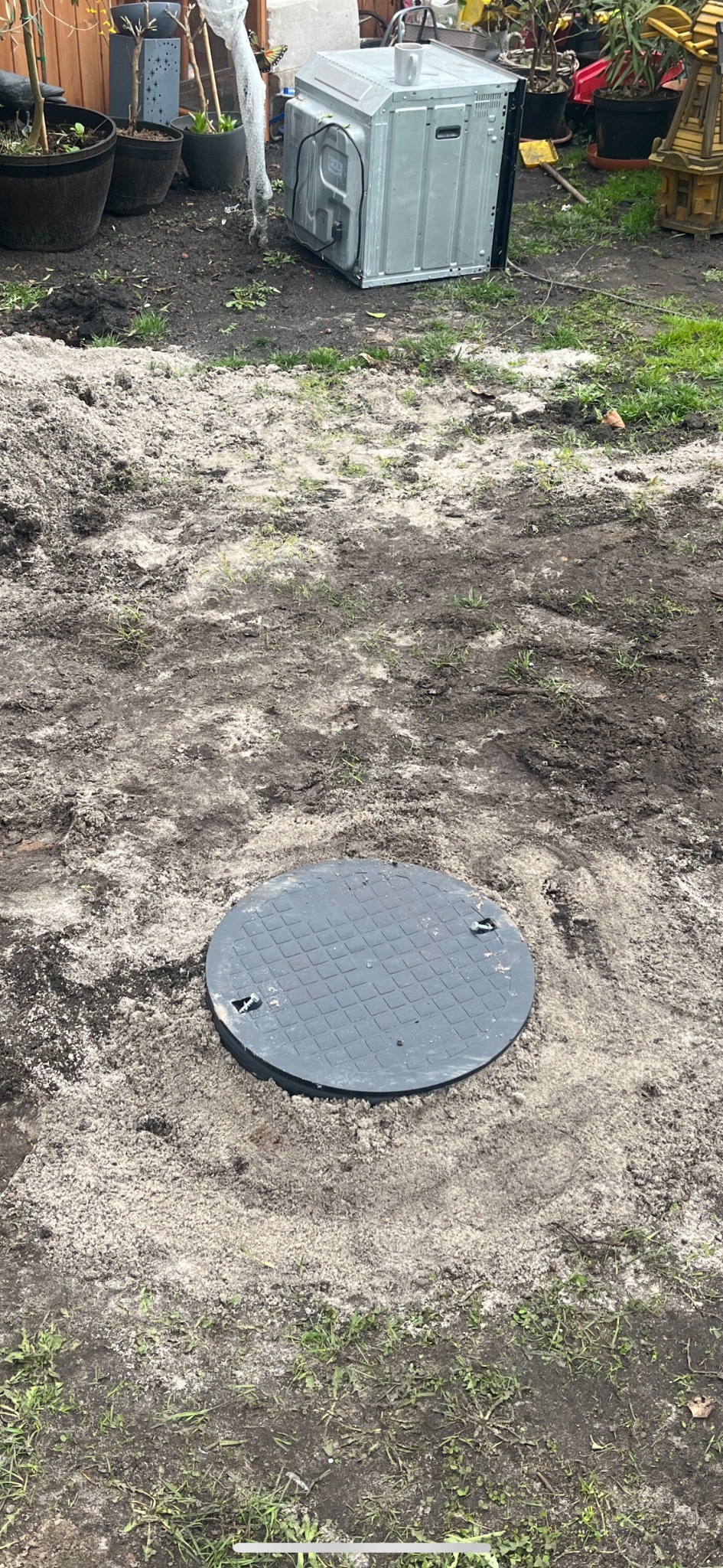 Okrągła, szara pokrywa studni na piaszczysto-ziemnym podłożu, z kuchenką w tle i elementami zieleni w ogrodzie.