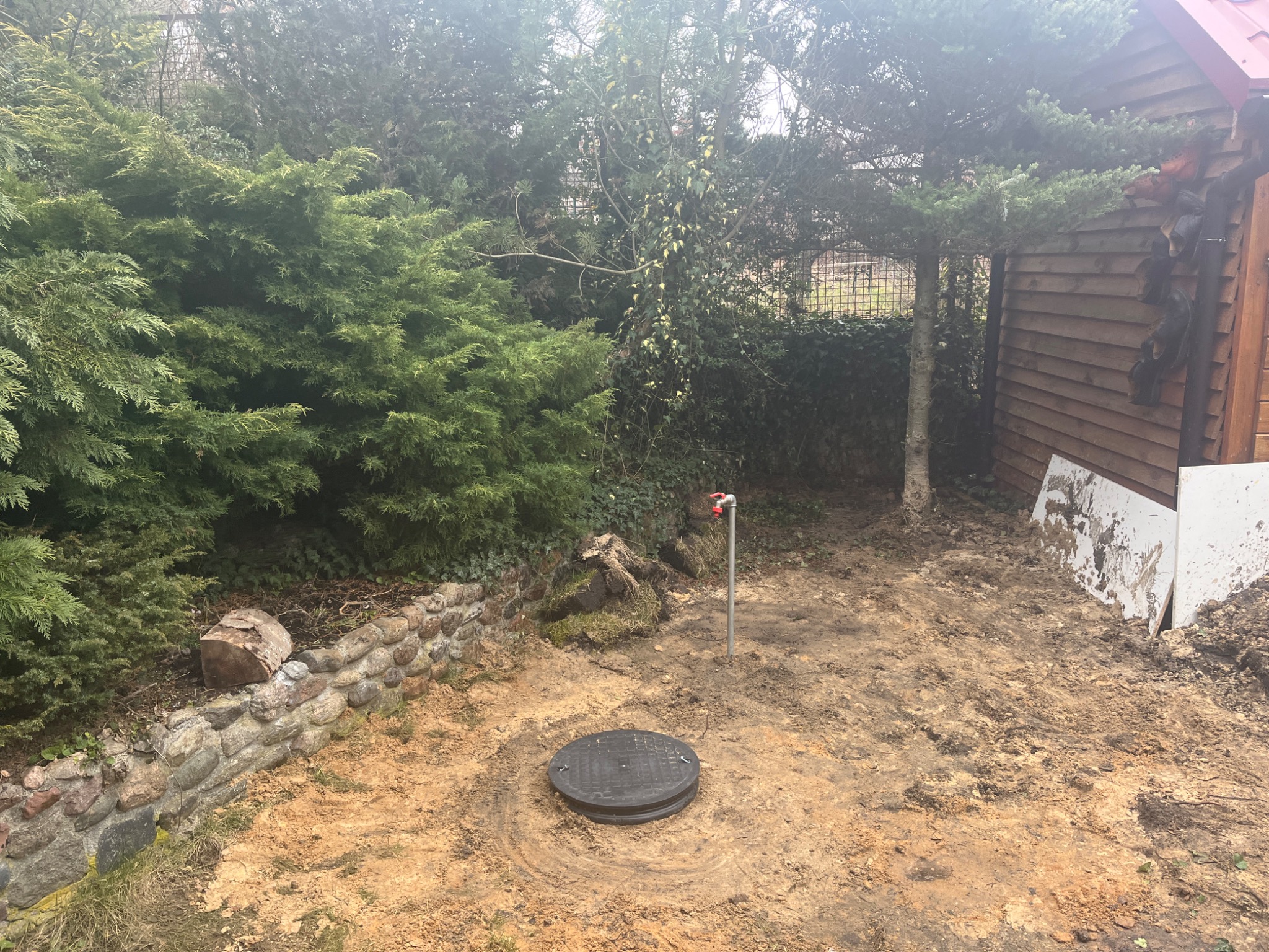 Otwór rewizyjny z czarną pokrywą w ziemi, obok mała instalacja z kranem, otoczone zielenią i drewnianym budynkiem.